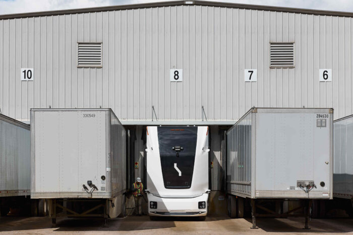 Ein weißer autonomer Elektro-Lkw von Einride steht an einer Laderampe zwischen zwei Aufliegern vor einem Logistikzentrum. Ein Mitarbeiter befindet sich neben dem Fahrzeug.